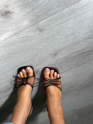 Bruna sandaler med mesh och rosett - Säljer ett par trendiga bruna sandaler med fyrkantig tå, tunna remmar i mesh och en söt rosettdetalj framtill. Perfekta för dig som gillar en stilren och modern look. Passar till både jeans och klänning.