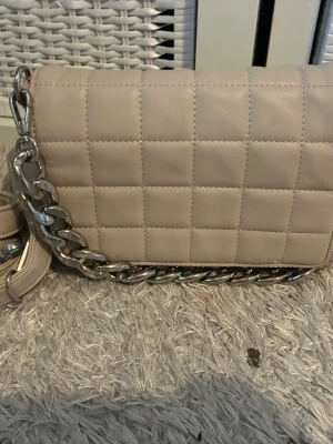 Beige quiltad axelväska med kedja - Säljer en beige axelväska med quiltat mönster och en rejäl silverfärgad kedja som axelrem. Väskan har en stilren, fyrkantig form och är gjord i ett material som liknar konstläder. Perfekt för dig som gillar trendiga accessoarer.