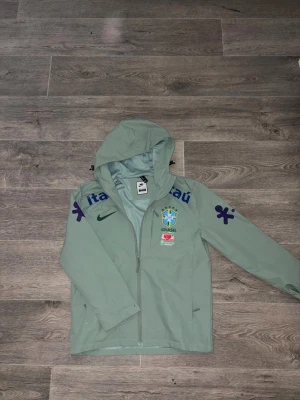 Nike Brazil Windbreaker - Ljusgrön vindjacka från Nike med Brasiliens landslagslogga och sponsortryck i blått. Jackan har huva, hel dragkedja och två sidofickor. Skickad samma dag! Skriv pm!!