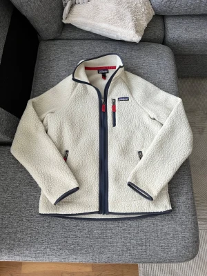 Vit fleecejacka från Patagonia - Säljer en vit fleecejacka från Patagonia med marinblå dragkedjor och röda detaljer. Jackan har hög krage, tre fickor med dragkedja och är tillverkad i mjuk, fluffig fleece. Perfekt för kyliga dagar och outdoor-häng.