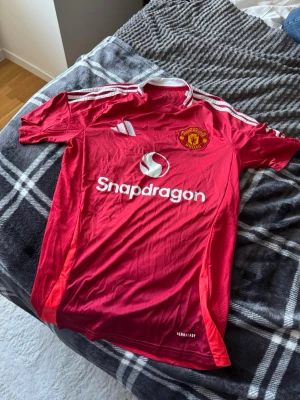 Manchester United matchtröja  - Röd Manchester United matchtröja från Adidas med vita detaljer och klubbmärke på bröstet. Tröjan har Snapdragons logga på framsidan och klassiska Adidas-ränder på axlarna. Tillverkad i lätt och ventilerande material, perfekt för fotboll eller träning.