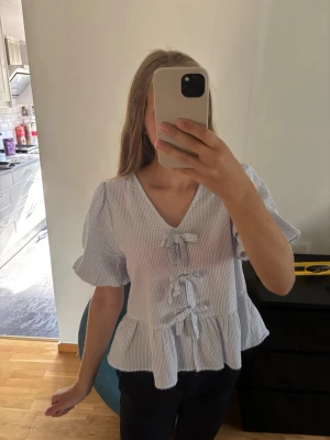 Vit- och blårandig blus med rosetter - Supersöt blus med tunna blå och vita ränder, v-ringning och tre dekorativa rosetter framtill. Blusen har korta puffärmar och volang nedtill. Aldrig använd. Storleken är 170 i barn men passar en tjej med storlek xs/s!