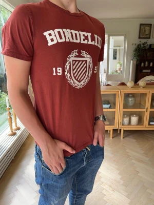 Bondelid T-shirt röd brun herr - Snygg Bondelid T-shirt i vinröd/burgundy färg med stort tryck framtill. Enkel att matcha och passar lika bra till jeans som chinos eller shorts. Perfekt vardagsplagg med klassisk college-inspirerad design.  #Bondelid #Tshirt #Burgundy #Vinröd #College #Preppy #OldMoney #Casual #Streetwear #Designer #Herr #Sommar #Trend #Mode #Basic #Logo #Kvalitet #Jeans #Chinos