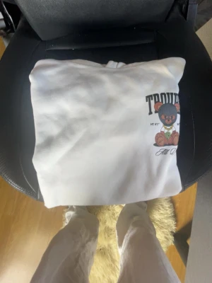 Vit hoodie med björntryck och text - Säljer en vit hoodie med stor tryckt björn på ryggen och texten 'TROUBLE'. Björnen har glasögon, guldhalsband och håller pengar. Hoodien har huva och ribbade muddar. Perfekt för dig som gillar streetwear och coola prints.