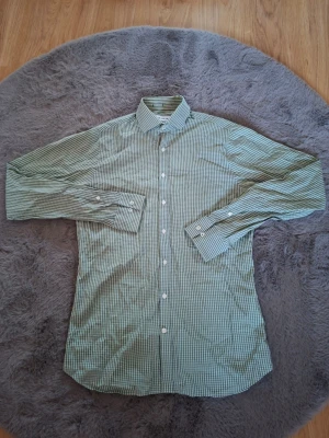 Grön rutig skjorta från The Shirt Factory - Snygg grön och blå rutig skjorta från The Shirt Factory i slim fit-modell. Skjortan har klassisk krage, långa ärmar med manschett och vita knappar framtill. Perfekt för dig som gillar stilrena och fräscha skjortor med lite smalare passform.
