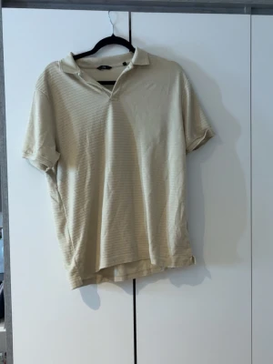 Beige pikétröja från C&A - Säljer en beige pikétröja från C&A med diskreta ränder och klassisk krage. Tröjan har korta ärmar och en relaxed passform, perfekt för en chill och enkel stil. Materialet känns mjukt och bekvämt mot huden.