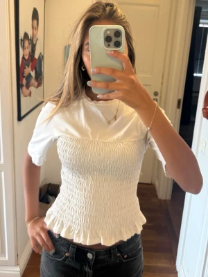 Vit smockad topp med volangkant - Supersöt vit topp med smockad midja och volangkant nedtill. Toppen har korta ärmar och rund halsringning. Perfekt att styla med jeans eller kjol för en trendig look. Materialet känns mjukt och stretchigt, vilket gör den bekväm att bära hela dagen.
