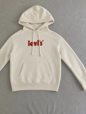 Vit hoodie från Levi's med orange logga - Snygg vit hoodie från Levi's med orange logga på bröstet. Hoodien har huva med dragsko och en stor magficka framtill. Tillverkad i mjukt bomullsmaterial som är skönt att ha på sig.