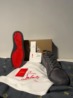 Gråa sneakers från Christian Louboutin - Snygga gråa sneakers från Christian Louboutin i mocka med klassisk röd sula. Skorna har lågt skaft, rund tå och matchande grå skosnören. Perfekta för dig som vill ha en exklusiv och stilren look med en ikonisk detalj under foten.