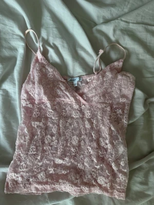 Ljusrosa spetslinnetopp - Säljer en ljusrosa linnetopp i spets med tunna axelband och v-ringad hals. Toppen är genombruten och har en feminin look som passar perfekt till sommaren. Superfin att styla med jeans eller kjol!