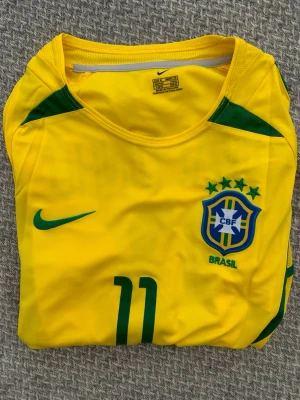 Brasilien fotbollströja Ronaldinho - Gul Brasilien fotbollströja från Nike med gröna detaljer, nummer 11 och Ronaldinho på ryggen. Tröjan har rund hals, korta ärmar och officiellt CBF-märke på bröstet. Perfekt för dig som älskar fotboll och vill stötta Brasilien.