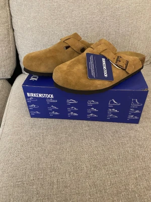 Bruna Birkenstock tofflor mocka 38 - Säljer ett par snygga bruna Birkenstock tofflor i mocka med justerbart spänne. Klassisk slip-in modell med mjuk fotbädd och robust sula. Perfekta för chill dagar hemma eller när du vill ha det bekvämt. Stl 38.