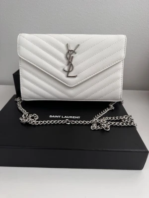 Vit kuvertväska  - Snygg vit kuvertväska med silverfärgad YSL-logga framtill. Väskan har quiltat mönster och en kedjerem i silvermetall. Tillverkad i ett tåligt material med elegant struktur och klassisk kuvertform.