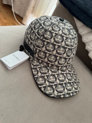 Mönstrad keps från Salvatore Ferragamo helt ny  - Säljer en snygg keps från Salvatore Ferragamo med beige och svart mönster över hela kepsen. Klassisk form med böjd skärm och justerbar rem bak. Tillverkad i ett slitstarkt tygmaterial och har exklusiv känsla. Perfekt för dig som vill sticka ut med stil.