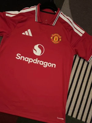 Manchester United Hemmaställ 26/27 Authentic - Säljer en röd Manchester United matchtröja från Adidas med vita detaljer och klubbmärke på bröstet. Tröjan har korta ärmar, vit krage och Snapdragons logga på framsidan. Perfekt för fotboll och träning.