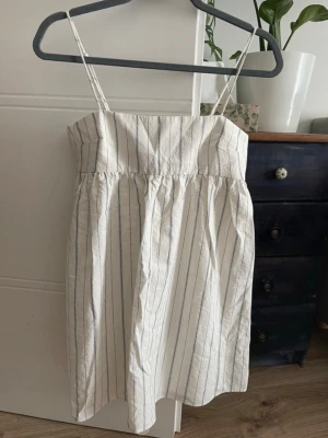 Randig sommarklänning med smala axelband med inbyggda shorts - Söt vit klänning med tunna blå ränder och smala axelband. Inbyggda shorts Klänningen har en rak överdel och lätt rynkad kjol för en avslappnad look. Perfekt för varma dagar och enkel att styla med sneakers eller sandaler.