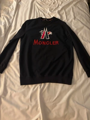 Svart Moncler sweatshirt med logga - Snygg svart sweatshirt från Moncler med stor broderad logga och text i rött, vitt och blått på bröstet. Tröjan har rund halsringning och långa ärmar. Perfekt för dig som gillar streetwear och vill ha något stilrent men ändå iögonfallande.