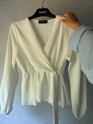 Vit omlottblus  - Snygg vit omlottblus från Missay med långa ärmar och knytband i midjan. Blusen har en lätt rynkad struktur och markerad midja som ger en feminin siluett. Perfekt att styla med jeans eller kjol för en trendig look.