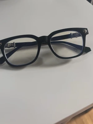 Svarta glasögon med silverdetaljer - Snygga glasögon med svart, fyrkantig båge och genomskinliga glas. Bågarna har coola silverdetaljer vid gångjärnen och ett diskret mönster på skalmarna. Perfekt accessoar för att lyfta din stil och ge en trendig touch till din look.
