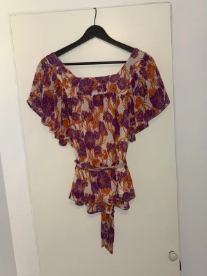 Blommig blus med volang och knytband - Supersöt blommig blus i lila och orange med volangärmar och knytband i midjan baktill. Blusen har en luftig passform och är perfekt för varma dagar.