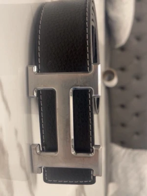 Svart skärp med silverfärgat H-spänne - Snyggt svart bälte i skinn med ett stort, silverfärgat H-format spänne. Bältet har vit kontrastsöm längs kanterna och en stilren, klassisk look som passar till många outfits. Perfekt accessoar för att lyfta din stil.