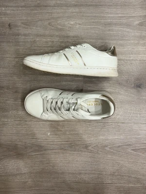 Vita sneakers med gulddetaljer - Snygga vita sneakers med guldfärgade detaljer och texten 'STOCKHOLM' på sidan. Skorna har klassisk låg profil, vita snören och guldfärgad hälkappa. Tillverkade i slätt läder för en clean look.