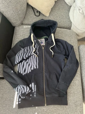 Mörkblå hoodie från Charles Wilson - Mörkblå zip-hoodie från Charles Wilson med huva och vita dragsnören. Klassisk modell med fickor framtill och ribbade muddar vid ärmslut och nederkant. Perfekt för en avslappnad stil.