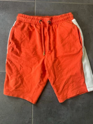 Orange shorts med vita sidor och snörning - Säljer ett par orange shorts med vita ränder längs sidorna och snörning i midjan. Shortsen har elastisk midja och två fickor fram. Perfekta för sommarens alla äventyr!
