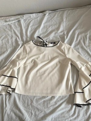 Vit blus med bell sleeves - Söt vit blus med bell sleeves, svarta detaljer i kanterna och knut i nacken. Tyget är smått genomskinligt.