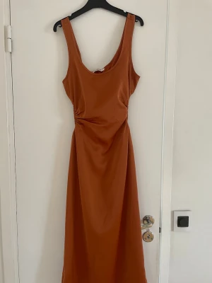 Klänning med öppen rygg - Snygg brun/orange långklänning i ett glansigt material med breda axelband och markerad midja. Klänningen har en öppen rygg med resårdetaljer och en enkel, elegant siluett. 