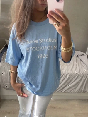 Blå oversized Acne Studios t-shirt - Säljer en ljusblå oversized t-shirt från Acne Studios med tryck 'STOCKHOLM 1996' framtill. T-shirten har korta ärmar, rund hals och är gjord i mjuk bomull. Perfekt för en chill och avslappnad stil.