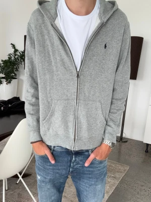 Grå zip hoodie från polo Ralph Lauren - Snygg gråmelerad hoodie med dragkedja och huva. Klassisk design med två stora fickor framtill och diskret logga på bröstet. Perfekt till jeans och enkel att matcha med andra plagg. Barn xl 18-20 men passar en mindre S