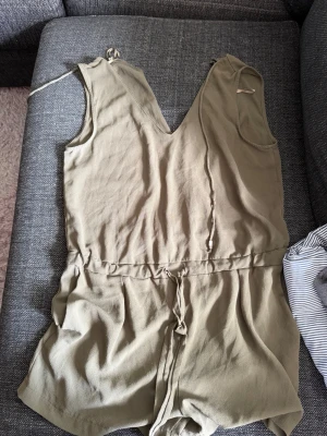 Beige kort jumpsuit med knytband - Snygg beige jumpsuit i mjukt, tunt tyg med korta ben och djup v-ringning både fram och bak. Midjan har ett knytband för att justera passformen. Perfekt för sommardagar och enkel att styla med accessoarer. Skriv privat för fler bilder eller om du har några frågor💗