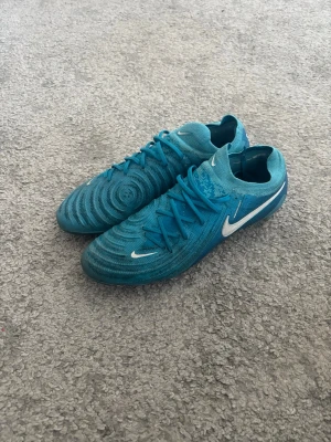 Blå Nike fotbollsskor med dobbar - Säljer ett par blå Nike fotbollsskor med vita detaljer och klassisk swoosh på sidan. Skorna har snörning och en strumpliknande passform för extra komfort. Ovandelen är i syntetmaterial och sulan har dobbar som passar för spel på konstgräs. Riktigt bra skick använda 4 gånger max