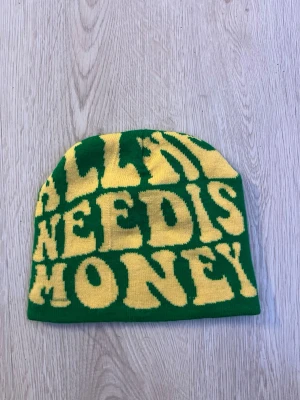Grön mössa med gul text - Säljer en grön mössa med stor gul text som säger 'ALL I NEED IS MONEY'. Mössan är stickad och har en avslappnad passform. Perfekt för dig som vill sticka ut med ett statement-plagg.