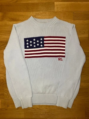 Ralph Lauren S - Ljusblå stickad tröja med ribbade muddar och krage. På bröstet finns en stor USA-flagga i rött, vitt och blått samt broderade initialer 'RL' i rött. Tröjan har lång ärm och klassisk passform. Perfekt för dig som gillar preppy stil.