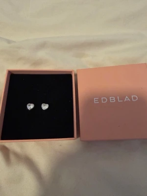 Hjärteformade örhängen från Edblad - Säljer ett par söta hjärtformade örhängen från Edblad i silverfärg. Örhängena har gnistrande stenar som ger en elegant touch och kommer i original ask. Perfekta för dig som gillar stilrena och klassiska accessoarer.