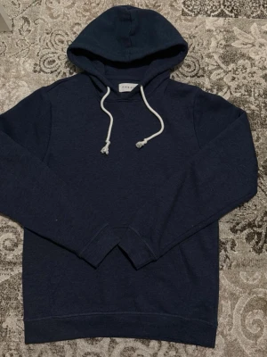 Mörkblå hoodie med vita snören - En stilren mörkblå hoodie med huva och vita dragsnören. Klassisk passform med långa ärmar och ribbade muddar. Perfekt för en avslappnad look och enkel att matcha med jeans eller joggers.