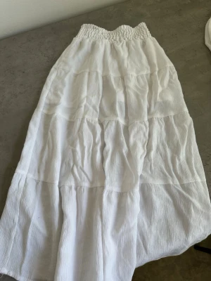 Vit långkjol med smock från H&M - Vit långkjol från H&M med smockad midja och mjukt, strukturerat tyg. Kjolen har flera sömmar som ger volym och en flowy känsla. Tillverkad i en blandning av bomull och polyester.