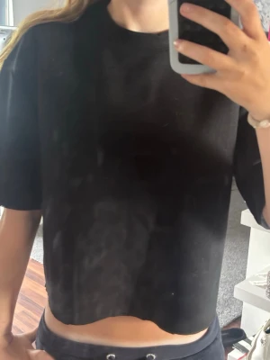 Svart croppad t-shirt - En enkel svart croppad t-shirt med rund hals och korta ärmar. T-shirten har en loose passform och är perfekt för en avslappnad stil. Materialet känns mjukt och bekvämt, troligtvis bomull eller en bomullsblandning.