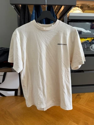 Beige t-shirt från Axel Arigato - Säljer en beige t-shirt i bomull med minimalistiskt svart tryck 'ARIGATO ©2014' på bröstet. Klassisk rund hals och korta ärmar. Perfekt basplagg för en clean och enkel stil.