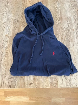 Mörkblå tunn hoodie från Polo Ralph Lauren - Mörkblå tunn hoodie från Polo Ralph Lauren med klassisk huva och dragsko. Tröjan har en liten röd broderad logga på bröstet och är tillverkad i mjukt bomullsmaterial. 