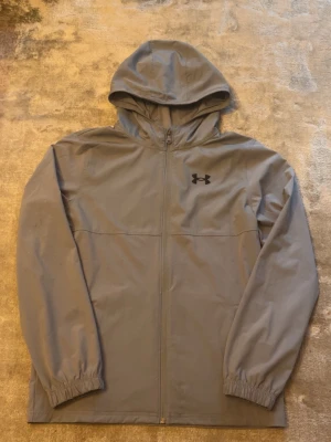 Grå vindjacka från Under Armour - Snygg grå vindjacka från Under Armour med huva och dragkedja framtill. Jackan har elastiska muddar vid ärmsluten och logga på bröstet samt text på ryggen. Tillverkad i lätt och vindtätt material, perfekt för blåsiga dagar.