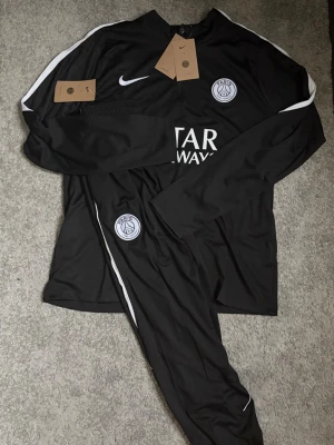 PSGTracksuit  - Säljer ett svart träningsset från PSG och Nike med långärmad tröja och matchande byxor. Setet har vita detaljer, PSG-logga på både tröja och byxa samt Nike swoosh. Materialet är lätt och andas, perfekt för träning eller uppvärmning.