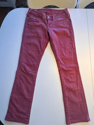Rosa/ Röda bootcut True Religion jeans - Säljer ett par rosa/röda true religon jeans med bootcut-modell . Byxorna har detaljerade sömmar och detaljerade bakfickor samt bälteshällor. Midjemått 33cm, ben innerlängd 74cm 