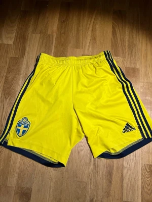 Gula Adidas fotbollsshorts Sverige - Säljer ett par gula fotbollsshorts från Adidas med blå detaljer och Sveriges landslagsemblem på ena benet. Shortsen har elastisk midja och klassiska Adidas-ränder längs sidorna. Perfekta för träning eller match!