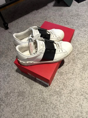 Valentino open - Snygga vita sneakers från Valentino med ett brett svart band över vristen och coola detaljer på sulan. Skorna har klassisk låg profil och är tillverkade i läder med vita snören och diskret logga på plösen. Perfekta för dig som vill ha en stilren men ändå unik look.