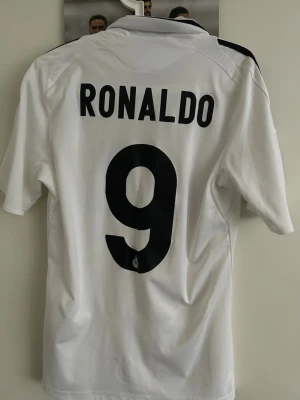 Real Madrid Ronaldo matchtröja - Säljer en vit Real Madrid matchtröja från Adidas med svarta detaljer och nummer 9 samt namnet Ronaldo på ryggen. Tröjan har klubbmärke, LFP-märke på ärmen och Bwin som sponsortryck på framsidan. Klassisk krage och korta ärmar.