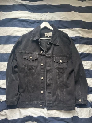 Svart jeansjacka Maison Margiela - Snygg svart jeansjacka från Maison Margiela med klassisk krage, vita knappar och två bröstfickor med lock. Jackan har raka ärmar och en avslappnad passform. Perfekt för dig som vill ha en stilren och tidlös look.