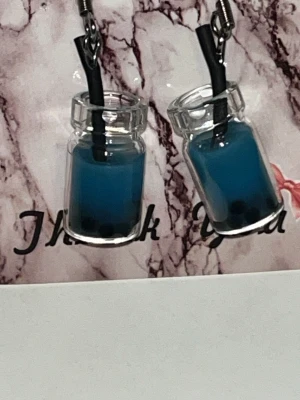Blå bubble tea örhängen i glas - Unika örhängen formade som små glasflaskor fyllda med blå vätska och svarta pärlor, inspirerade av bubble tea. Örhängena har svarta sugrörsdetaljer och hängs på krokar i metall. Perfekta för dig som vill sticka ut med en lekfull accessoar. Örhängens krokarna är i rostfritt stål
